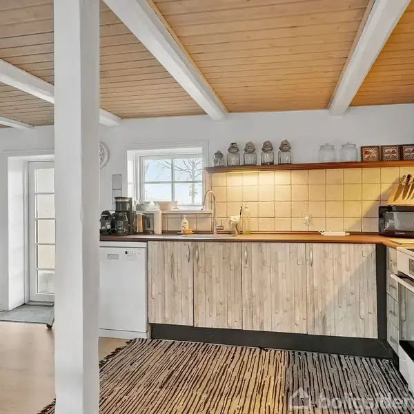 Køkken med træpaneler og moderne hvidevarer, der er placeret mod en flisebelagt væg. En tæppebeklædt gulvflade pryder området i et lyst rum med synlige loftsbjælker.