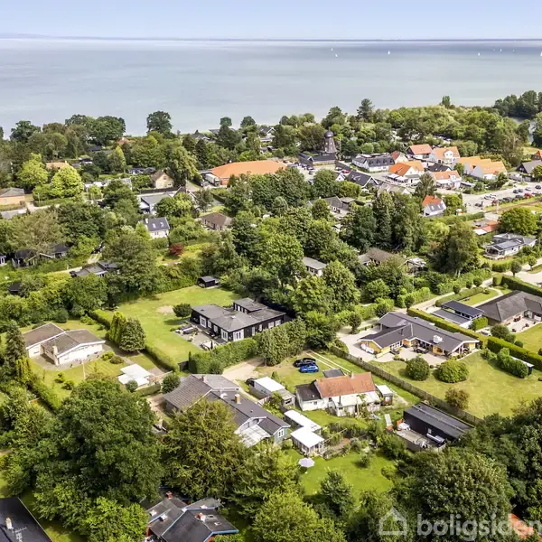 Bebyggelse set oppefra; huse er omgivet af træer og haver. I baggrunden strækker havet sig ud mod horisonten, og flere huse danner et landsbymiljø.