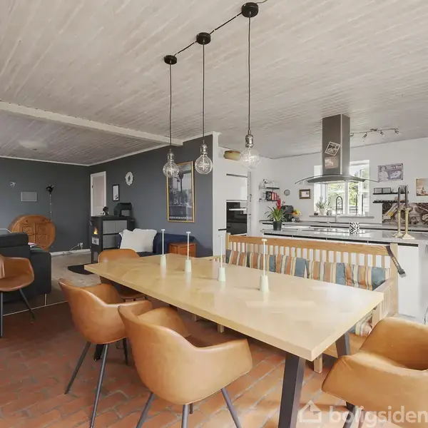 Et lille grønt træhus med hvidrammede vinduer står fredeligt ved en grusvej. I haven er der et træ, og en opslagstavle med teksten "Køn lille helårsbungalow".
