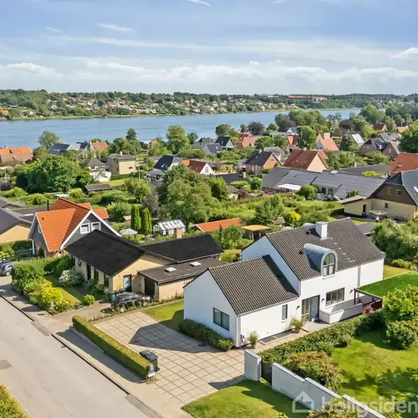 Flere lyse og mørke tage huse står stille i en grøn beboelseskvarter tæt på en sø med skovfyldte bakker i baggrunden under en blå himmel.
