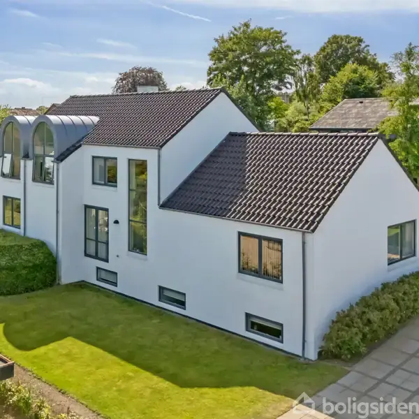 Et hvidt, moderne hus med sorte tagsten står omgivet af en velplejet have og terrasse. Store vinduer præger facaden, mens buske omkranser ejendommen. Solrig himmel skaber en lys atmosfære.