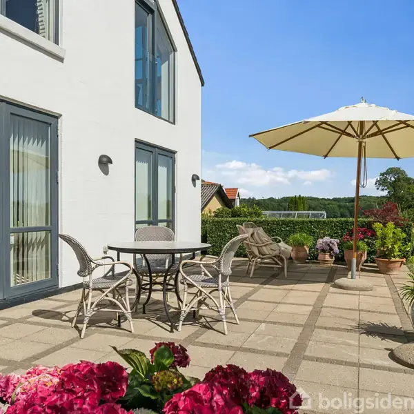 Hvid bygning med store vinduer, terrasse med havemøbler og parasol. Solrig have med grønne planter og blomster, omgivet af natur.