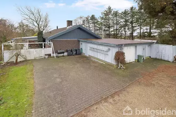 Et parcelhus med garage og indkørsel. Huset har murstensvægge og er omgivet af træer og en græsplæne. I indkørslen står flere skraldespande op ad en hvid væg.