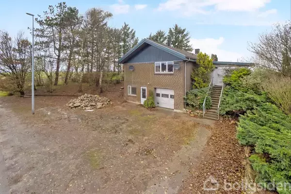 Et hus står i et skovklædt område med en garage i stueetagen. En trappe fører op til hovedindgangen med buske langs stien. En lysning omgiver huset.