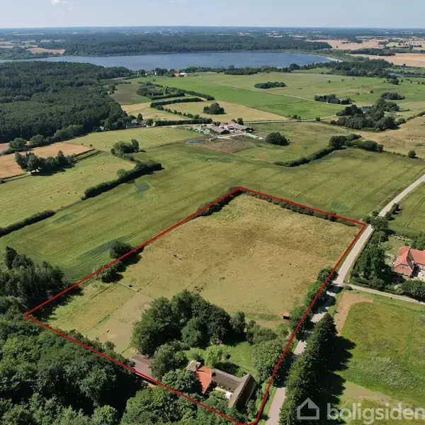 En stor, markeret grund ligger i et frodigt, landligt område med marker og skov, omkranset af veje. I baggrunden ses en sø.