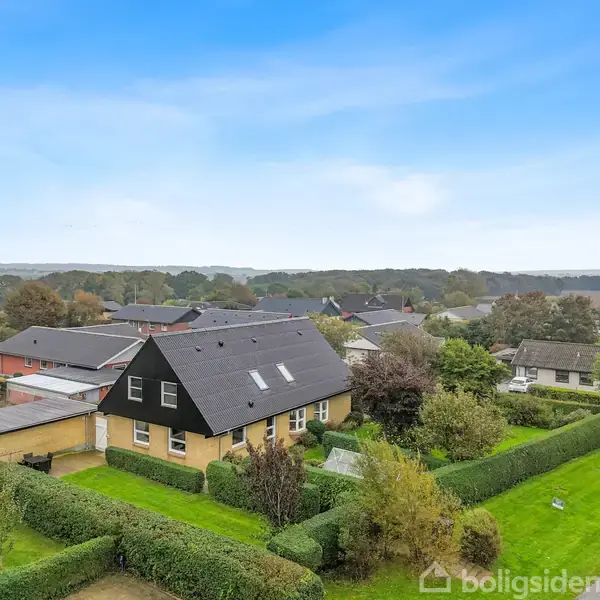 En moderniseret gul villa med sort tag og solpaneler står omgivet af en velplejet have og grønne buske. Huset befinder sig i et roligt kvarter med flere lignende boliger.