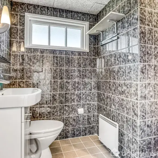 Vask står stationært ved toilettet i et badeværelse med sorte, blanke fliser; et vindue lader lys trænge ind. Vægbelysning over vasken, og bruser ved siden af.