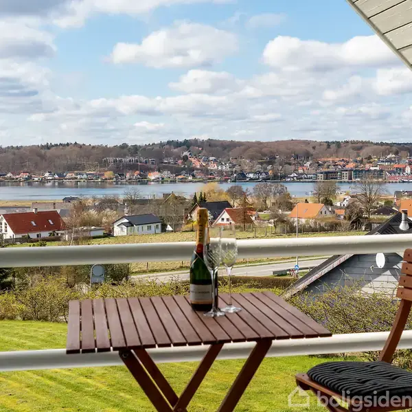 Et lille bord med en flaske champagne og to glas står på en altan. I baggrunden ses en grøn have og en idyllisk udsigt over vand og landsby.