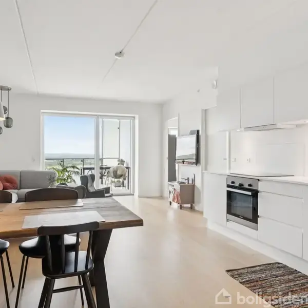 En moderne stue og køkken i et åbent rum. Et spisebord med stole står foran et lyst køkken med indbyggede hvidevarer. Sofaen fylder ved vinduet med balkonudsigt.