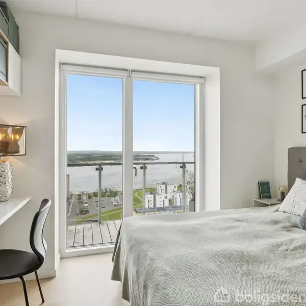 Soveværelse med dobbeltseng stående ved siden af en glasdør, der fører til en balkon med udsigt over en flod og bygninger. Et skrivebord med lampe står ved siden af en stol.