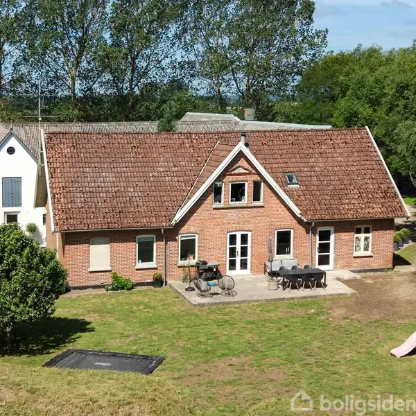 Rødstenshus med terrasse og havemøbler, beliggende i en grøn have med træer og en legeplads. Ved siden af står en hvid bygning.