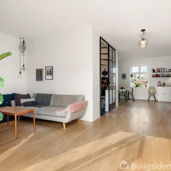 En stue med en grøn plante og sofa til venstre, et skrivebord og hylder i baggrunden. Rummet har trægulve og lyse vægge, med moderne indretning og belysning.