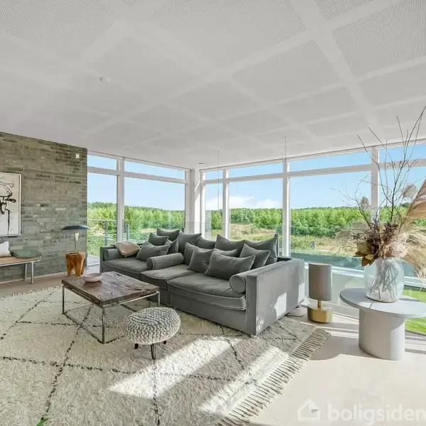 En rummelig stue med en stor, grå sofa står ved en væg af vinduer. Et tæppe ligger på gulvet, og der er et sofabord foran sofaen. Udenfor ses grønne træer.