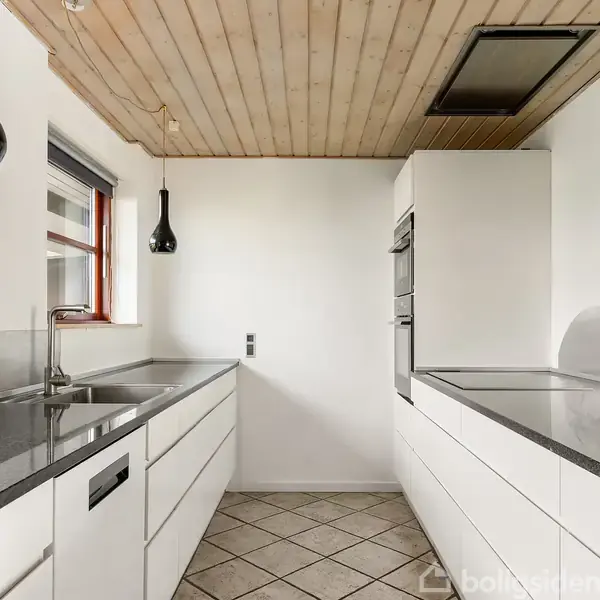 Et moderne køkken med hvide skabe har en skinnende grå bordplade. To hængelamper, i sort og rød, hænger fra det lyse træloft. Gulvet har kvadratiske fliser.