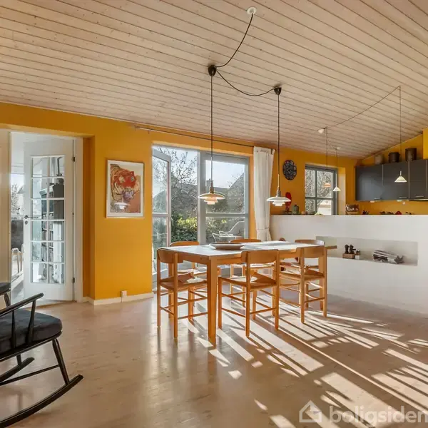 Et lyst køkkenalrum med spisestuemøbler i træ. Lyse træpaneler på loftet, en gyngestol, hvide skabe, og store vinduer lader sollys skinne ind. Gul vægfarve skaber en varm atmosfære.