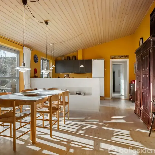 Et lyst køkken-alrum med gule vægge har et spisebord med stole i forgrunden. Tre pendellamper hænger over bordet. I baggrunden ses et åbent køkken og en glasdør til terrasseområdet.