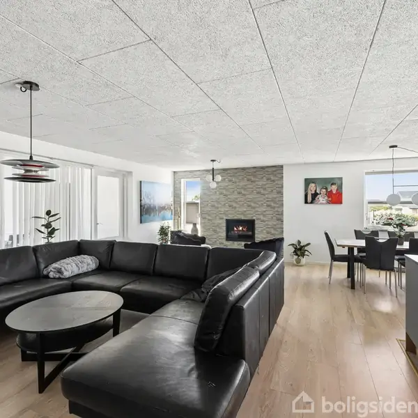 Sort lædersofa formet som en L, står ved et rundt sofabord i en moderne stue. Stuen har lyse lofter, parketgulv, spisebord med sorte stole, pejs og dekorationer.