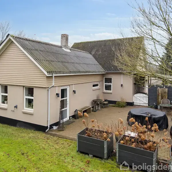 Et enetages hus med skråt tag står i en have. Foran huset er en terrasse med udendørs møbler og plantekasser. Omkring ses græsplæne og træer.