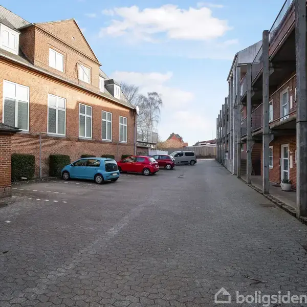 Flere parkerede biler står stille på en brostensbelagt gårdsplads mellem to rødstensbygninger under en blå himmel med skyer.