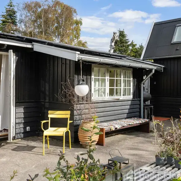 Et sortmalet, enkelt sommerhus med åben dør til terrasse, hvor der står en gul stol og en bænk ved siden af planter. Omgivelserne er grønne træer under en blå himmel.