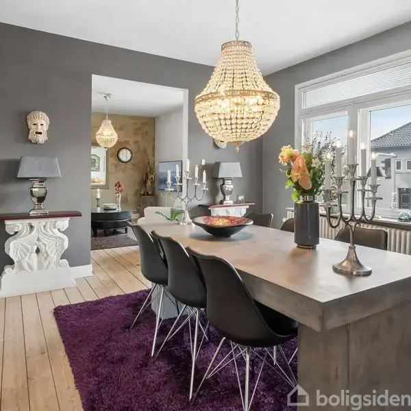 Et stort spisebord med sorte stole står på et lilla tæppe. Ovenover hænger en stor lysekrone. Væggen er grå, og der står en dekorativ lampe og maske på et konsolbord.