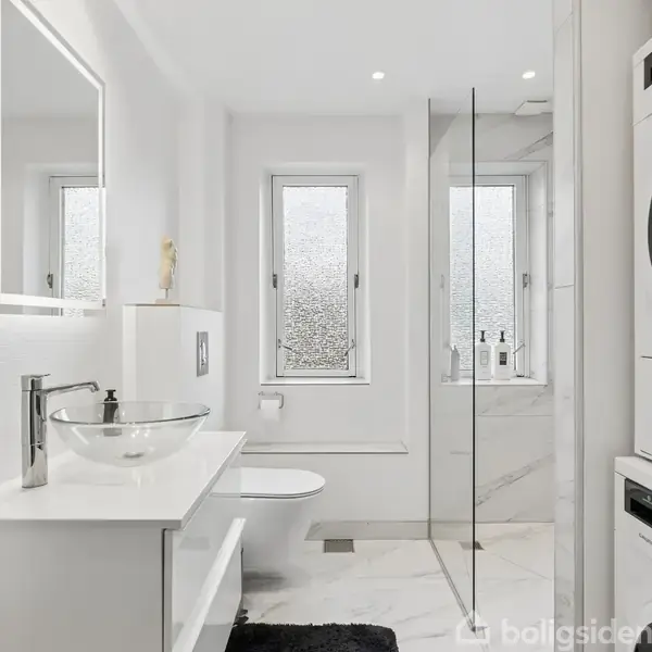 Et moderne badeværelse med en glasvask på et hvidt skab, en vægmonteret toiletkumme, og en brusekabine med glasvæg. Der er en vaskemaskine og tørretumbler stablet i hjørnet.