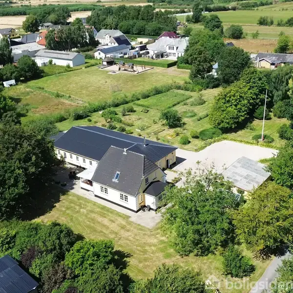 Et større hus med mørkt tag omgivet af grønne træer i et landsbyområde. Omkring ejendommen er der haver og græsplæner. I baggrunden ses flere huse og åbne marker.