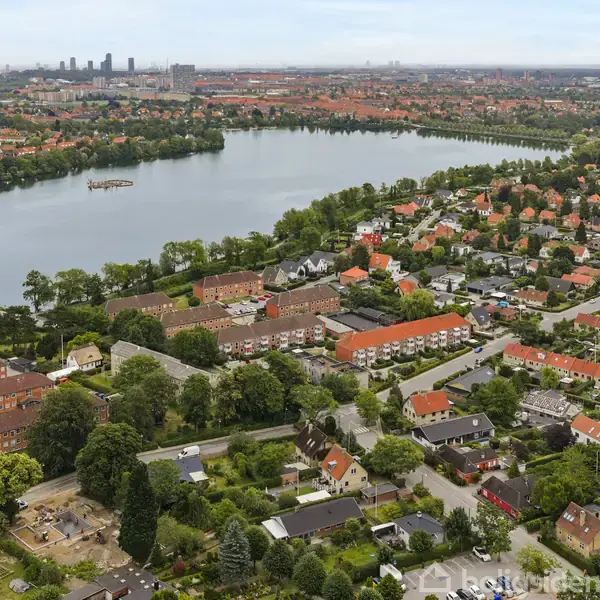 En sø omgivet af boligkvarterer med røde tage, træer langs søens bredder, og byens skyline i baggrunden.