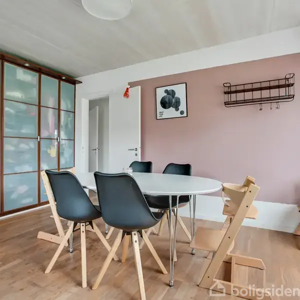 Et rundt bord med sorte stole omkring står i et lyst rum med trægulv. En rosa væg har kunst og en hylde. Skydedøre i glas til venstre.