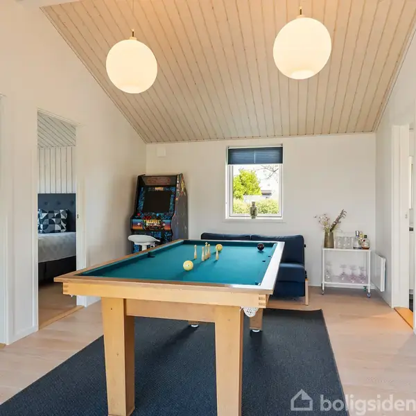 Et billardbord står centralt i et lyst værelse med træloft. En flippermaskine står i hjørnet, mens en sofa og et lille bord ses ved vinduet.