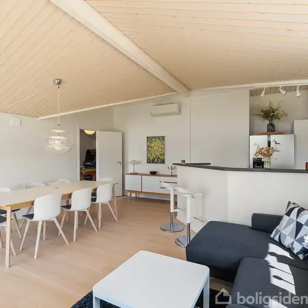 Stue med spisebord og stole til højre, en sort hjørnesofa med pude til venstre, åben forbindelse til et lyst køkken, hængende lampe og TV på væggen.
