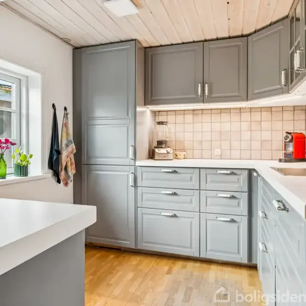 Gråt køkken med lyse træborde og skabe i L-form; kande og kaffemaskine på bordet. Blomster og viskestykker ved vindue. Udsigt over gårdhave. Indbydende og lys atmosfære.