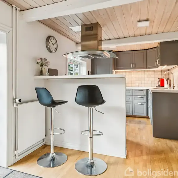 To høje barstole står ved en hvid køkkenø i et lyst køkken. Bag dem ses grå skabe, en vægur og et vindue til en terrasse med græsplæne udenfor.