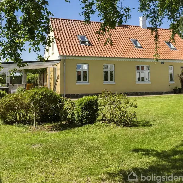 Et gult hus med rødt tegltag står stille i en velplejet have med grønne træer og buske. En solrig dag i et frodigt område.
