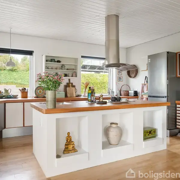 Et moderne køkken med en central køkkenø dekoreret med vaser og en Buddha-figur. Køleskabet står langs væggen, og store vinduer giver udsigt til en grøn have.