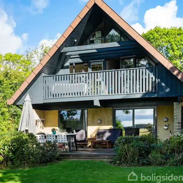 Et A-formet hus med to etager står i en frodig have. Øverste etage har en balkon, og nederst ses en terrassen med møbler og parasol.