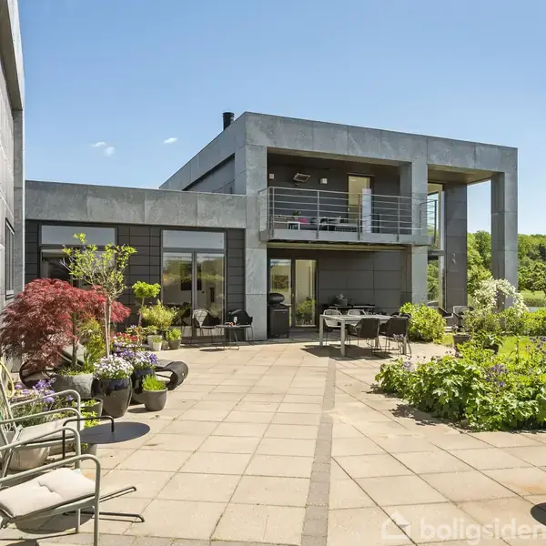 Moderne hus med store vinduer og en altan. Terrasse fyldt med havemøbler og planter. Grønt område i baggrunden under en klar blå himmel.