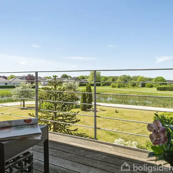 En terrasse med et bord og planter fremviser en udsigt over en grøn eng med træer og en sø i baggrunden under en klar blå himmel.