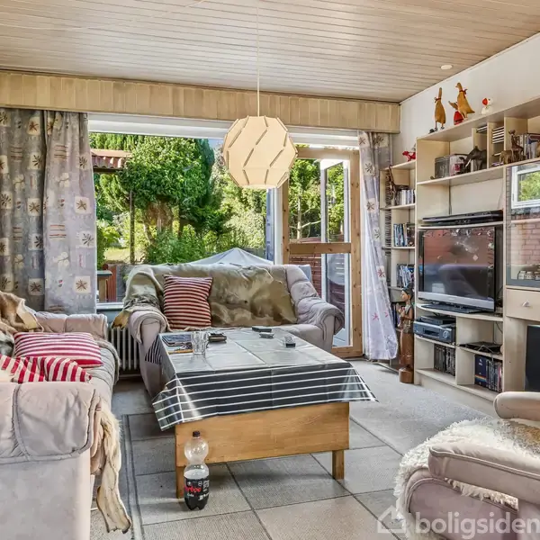 Stuen har en sofa og et sofabord med stribede puder. Der er en åben dør til en terrasse med haveudsigt. En reol indeholder en TV-skærm og dekorationer.