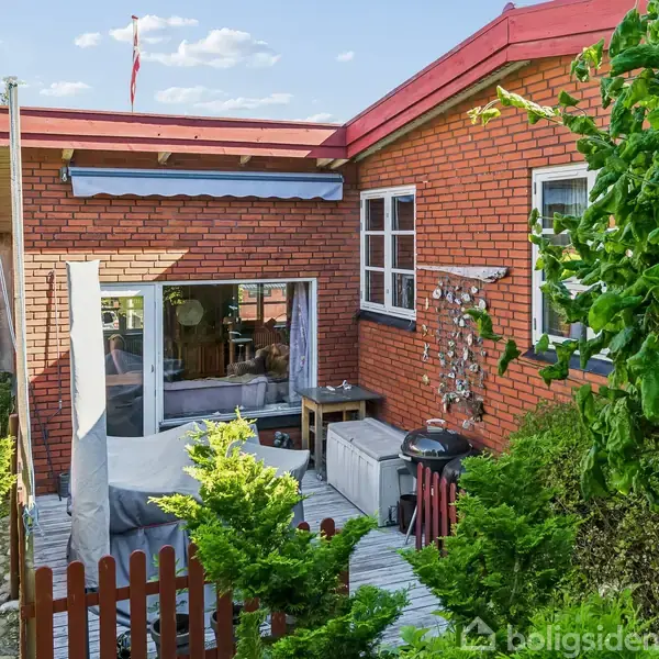 Rød murstenshus med store vinduer viser en udendørs terrasse med møbler. Grønne planter omgiver området, og en dansk flagstang står ved siden af huset under en blå himmel.