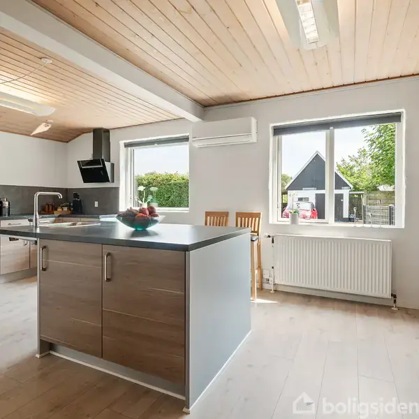 Køkken med en centreret ø. Køkkenøen har skuffer; ved vinduerne ses spisestole. Moderne apparater er integreret, to vinduer giver lysindfald, og trægulv omkranser.