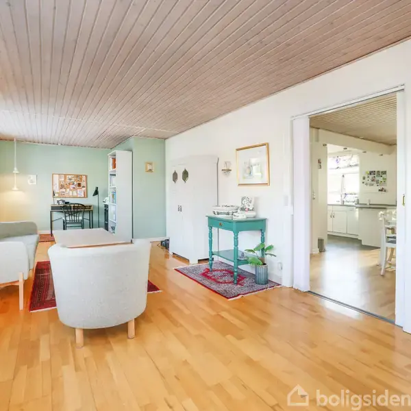 To stole står på et trægulv i en lys stue, der er forbundet til et køkken. Billeder og planter dekorerer væggene og et lille bord.