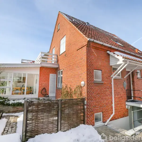 Rødt murstenshus med skråt tag, snebelagt have og stenbelægning. Vedhæftet er en glasudbygning til venstre. Skyfri himmel danner en lys og åben atmosfære.