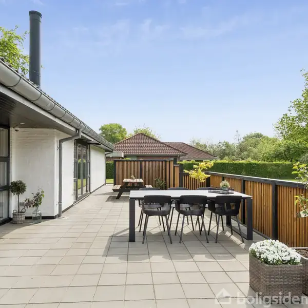 En moderne terrasse med bord og stole står ved et hus. Omgivet af grønne træer og græsplæne, udgør det en moderne udendørs spisestue. Der er ingen tekst i billedet.