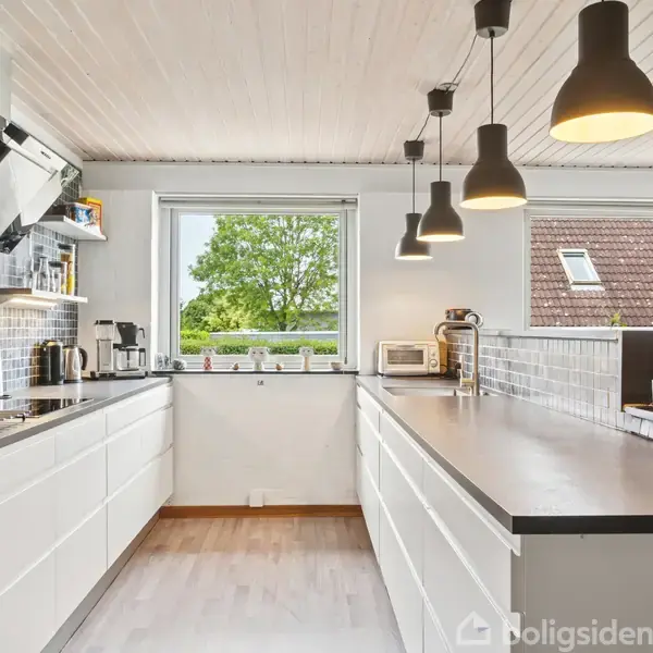 Køkken med hvide skabe står i et L-layout. Moderne lamper hænger fra loftet. Et vindue viser grønne træer udenfor. Kaffemaskine og brødrister står på bordet.