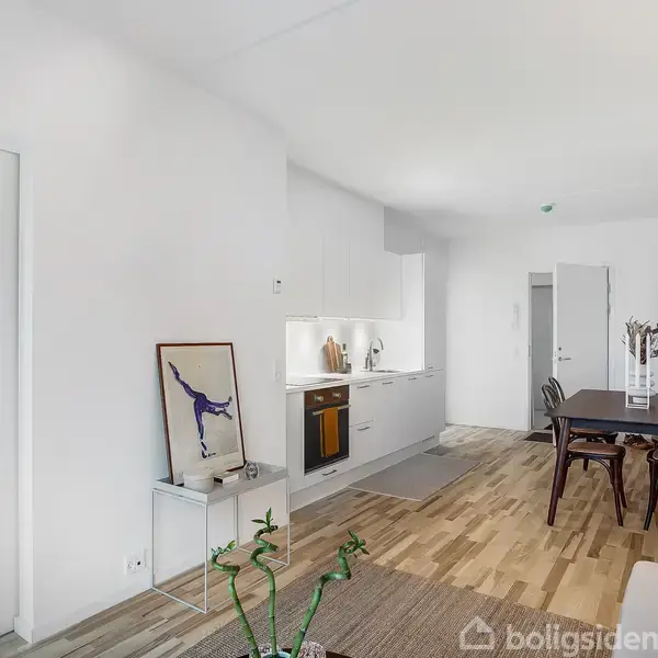 Stue med hvidt køkken langs væggen, spisebord med stole til højre. En åben dør fører til et lyst værelse. Moderne indretning med lyst træ og sofa.