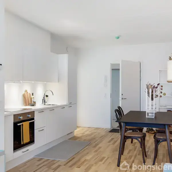 Et moderne køkken med indbyggede hvide skabe og en ovn, der er placeret ved siden af en spisekrog med et sort bord og fire stole. Lyse trægulve og hvidmalede vægge.