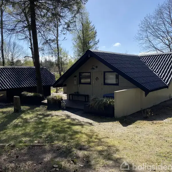 Et sommerhus med sort tag ligger i en skovklædt have. Ved siden af ses en mindre bygning. Træer omkranser området, hvilket skaber en fredfyldt og naturskøn atmosfære.