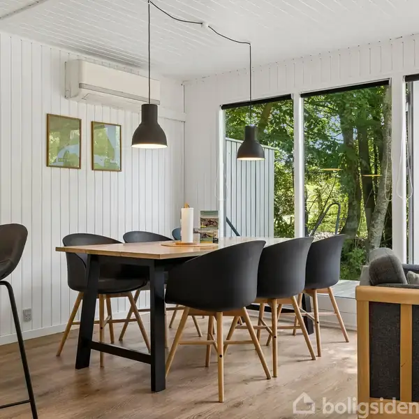 Stue med grå sofaer og brændeovn, åbner mod en have via store vinduer. Et spisebord med stole står i baggrunden, og interiøret er lyst med træloft og -gulv.