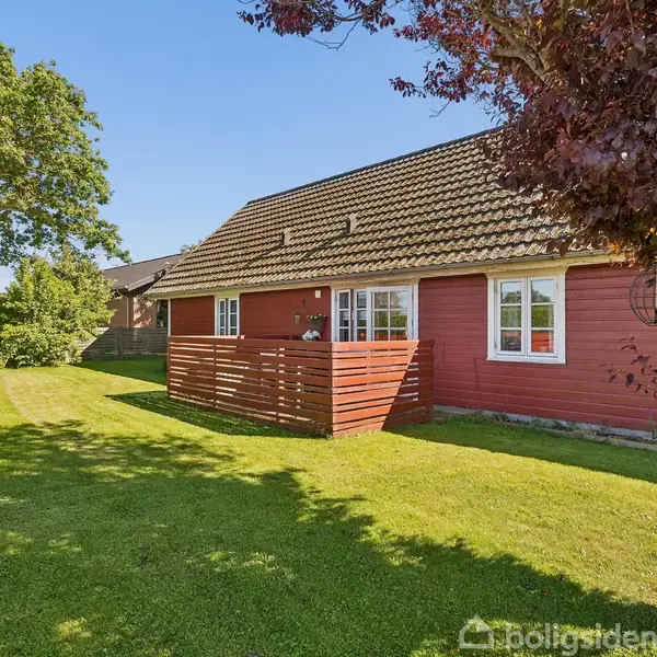 Rødt hus med hvide vinduer står stille i en solrig have med grøn græsplæne og store træer. En lille træterrasse er foran huset.